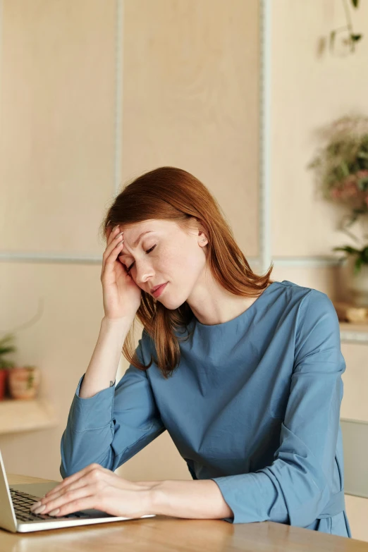 a woman sitting at a table with a laptop, two exhausted, wearing a light blue shirt, showing forehead, sleep deprived