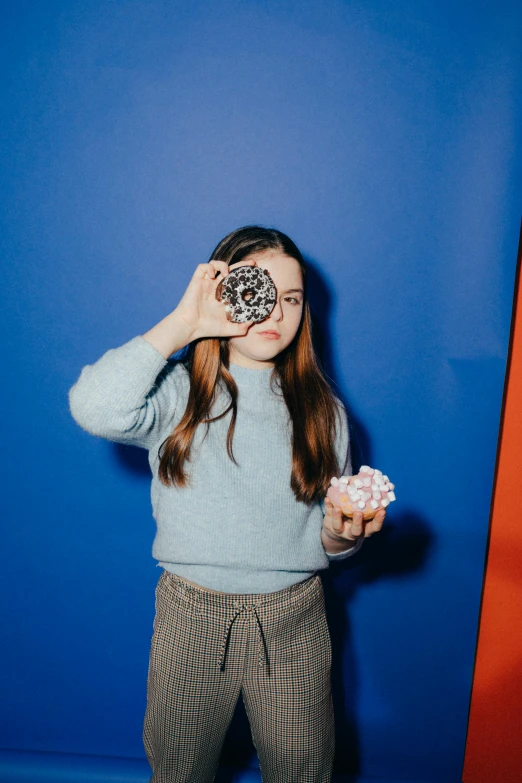 a woman standing in front of a blue wall holding a donut, pexels contest winner, op art, maisie williams, mackenzie foy, holding a camera, is looking at the camera