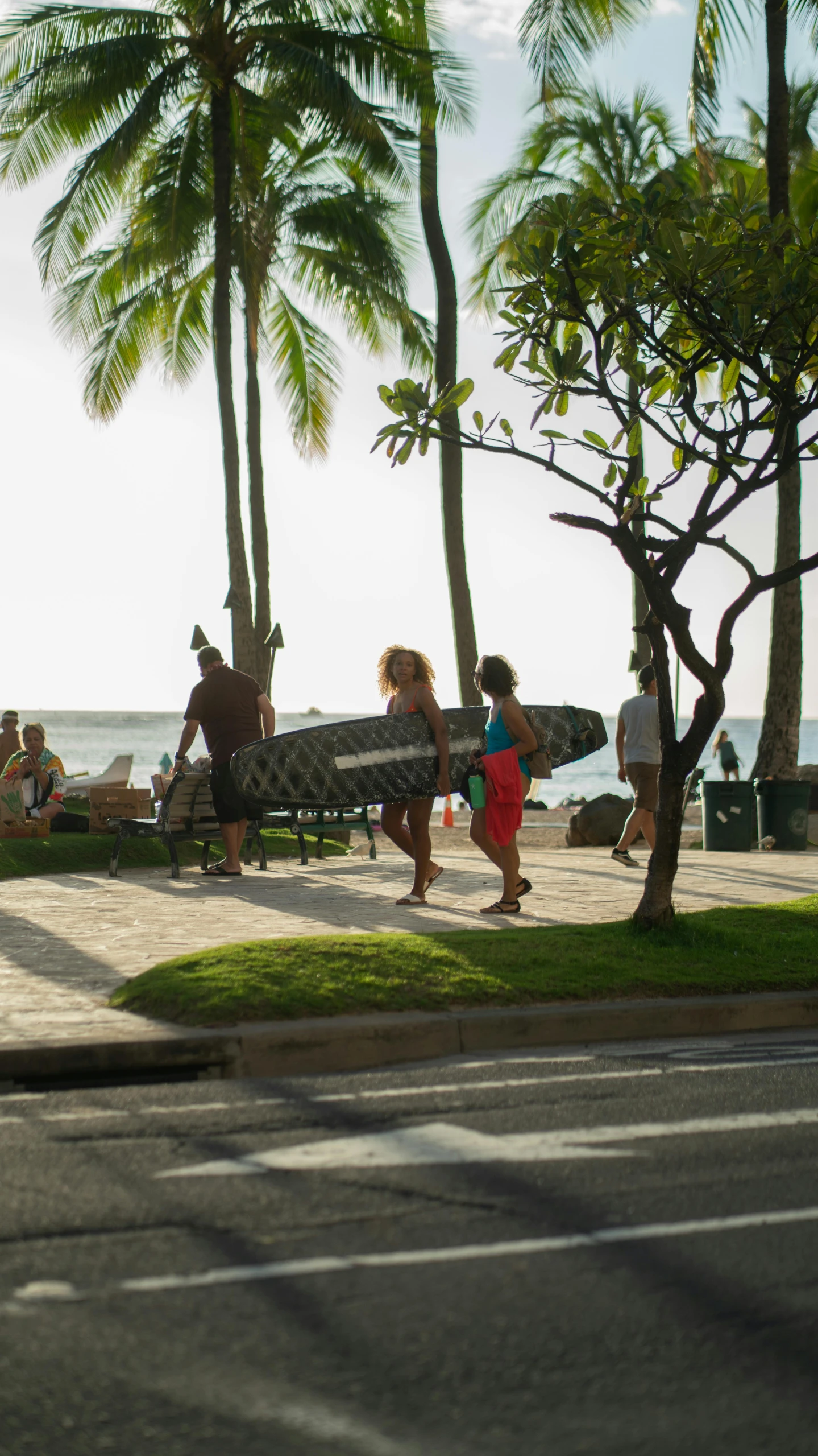 a group of people walking down a sidewalk next to the ocean, standing on surfboards, waikiki beach, bench, square