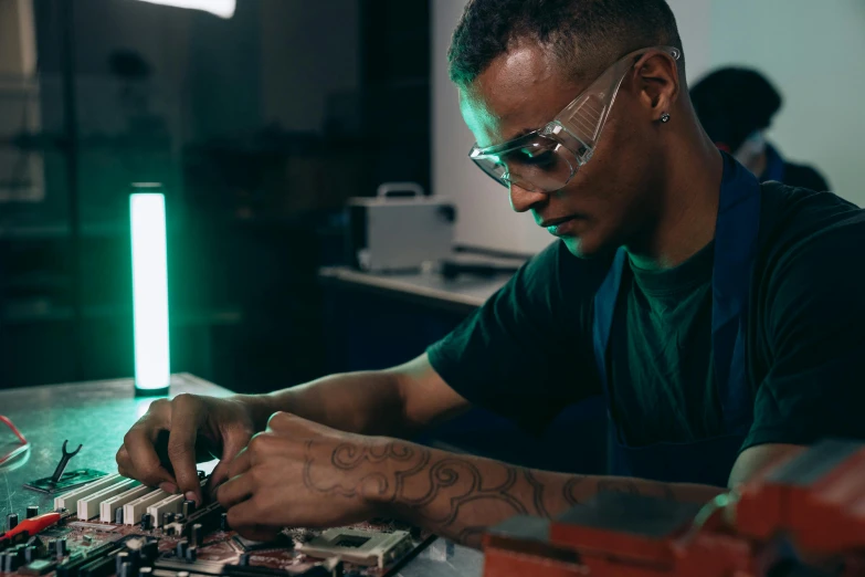 a man sitting at a table working on a computer, an etching, trending on pexels, afrofuturism, circuit boards, artist wearing overalls, wearing black rimmed glasses, professional gunsmithing