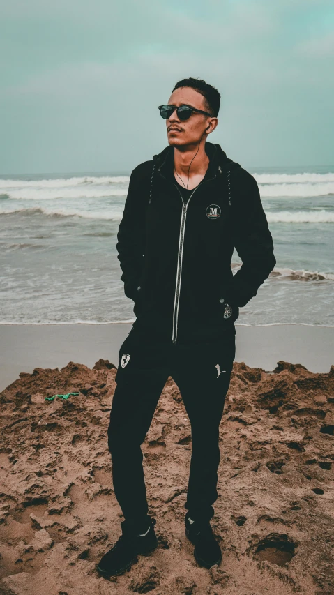 a man standing on a beach next to the ocean, a cartoon, by Robbie Trevino, pexels contest winner, dressed black hoodie, wearing a track suit, portrait mode photo, full body-n 9