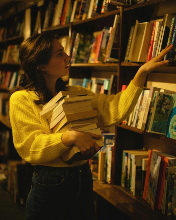 a woman holding a stack of books in front of a bookshelf, a screenshot, happening, lesbians, analogue photo low quality, on a yellow canva, early evening