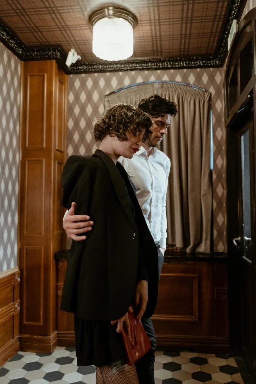 a man standing next to a woman in a room, inspired by Nan Goldin, pexels, renaissance, two men hugging, finn wolfhard, ornate back tuxedo, ( ( theatrical ) )