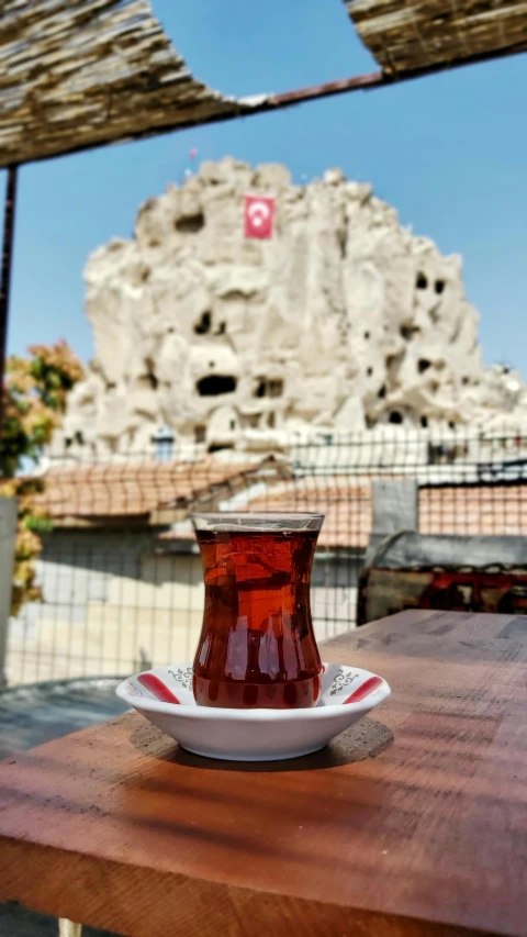 a cup of tea sitting on top of a wooden table, by irakli nadar, temple in the background, stone grotto in the center, iced tea glass, can basdogan