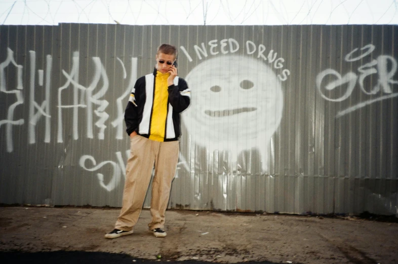 a man standing in front of a wall with graffiti on it, an album cover, inspired by Nick Fudge, graffiti, mustard, outside alone smoking weed, wearing a track suit, california