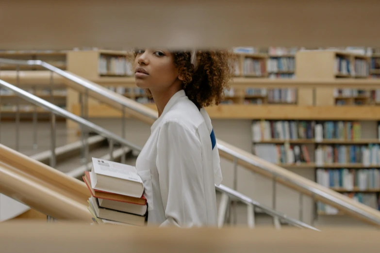 a woman holding a stack of books in a library, pexels contest winner, with brown skin, walking to the right, wearing lab coat and a blouse, black teenage girl