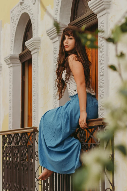 a woman sitting on a balcony next to a building, by Alejandro Obregón, pexels contest winner, long skirt, long hair with full bangs, warm azure tones, promotional image