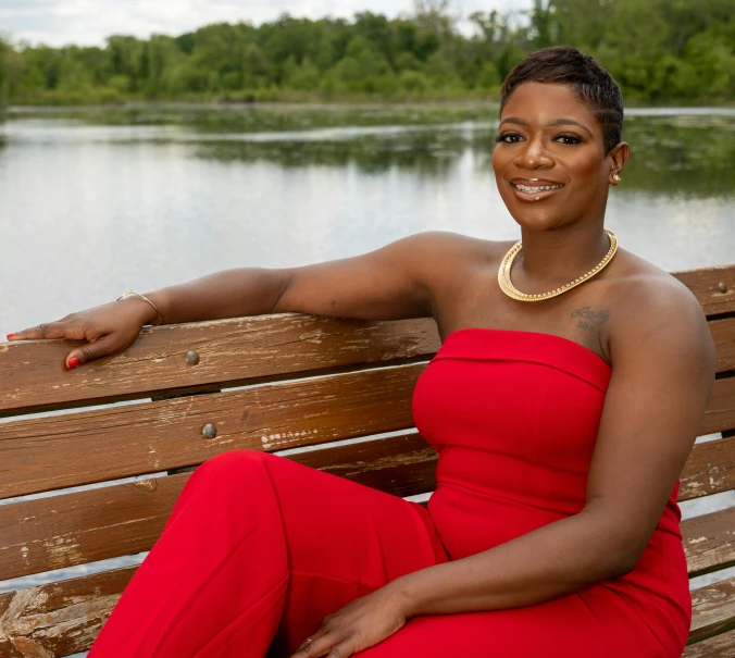 a woman in a red dress sitting on a bench, photo of a black woman, lake setting, wearing red formal attire, lgbtq