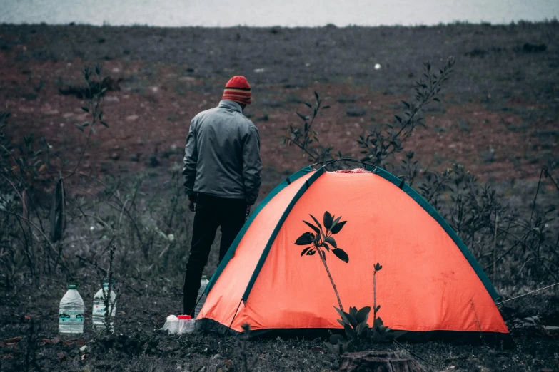 a man standing next to a tent in a field, pexels contest winner, 🚿🗝📝, bright orange camp fire, post grunge, bushes in the background