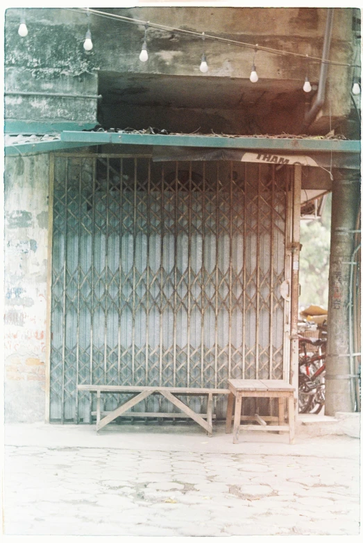 a person sitting on a bench in front of a building, iron arc gate door texture, 1970s philippines, shop front, cell bars