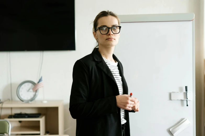 a woman standing in front of a white board, by Adam Marczyński, pexels, academic art, in square-rimmed glasses, wearing a black jacket, teacher, looking serious