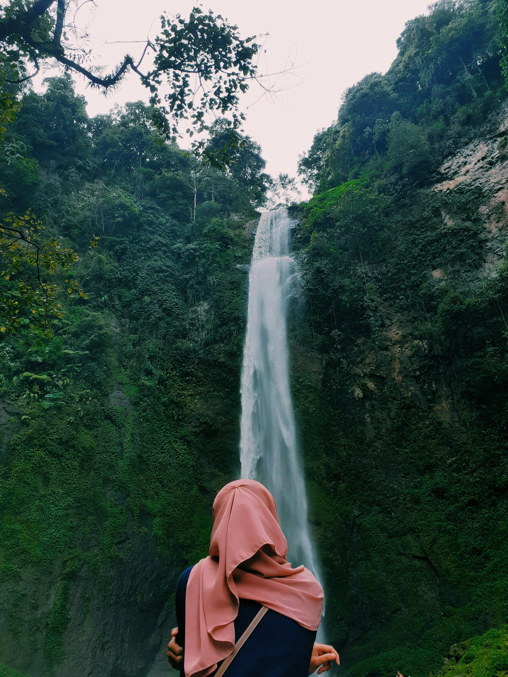 a woman standing in front of a waterfall, by Basuki Abdullah, towering high up over your view, hijab, from the distance, low quality photo