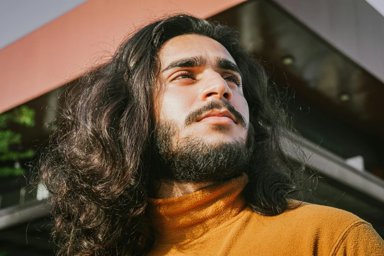 a man with long hair standing in front of a building, a character portrait, by Adam Dario Keel, trending on pexels, middle eastern skin, wavy hair yellow theme, square masculine jaw, wearing turtleneck