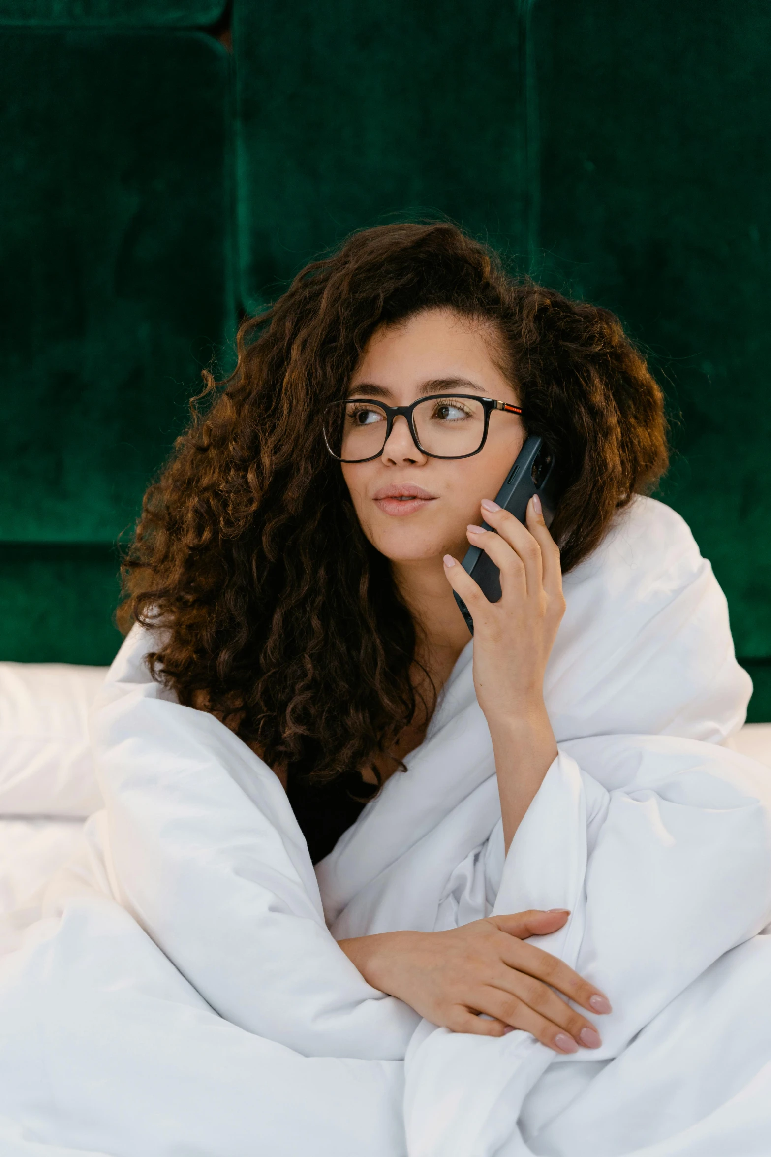 a woman laying in bed talking on a cell phone, trending on pexels, renaissance, wavy long black hair and glasses, tech robes, curly brown hair, pondering