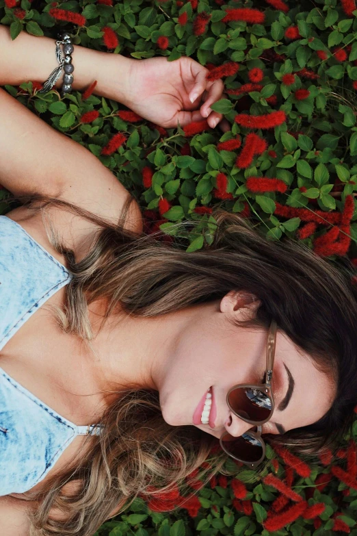 a beautiful young woman laying on top of a lush green field of flowers, inspired by Elsa Bleda, trending on pexels, with sunglass, green and red plants, smiling down from above, chloe bennet