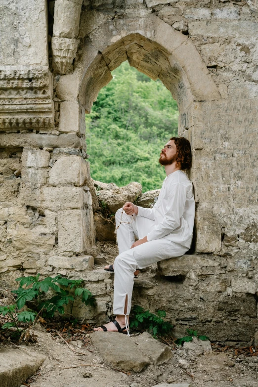 a man sitting on top of a stone wall, an album cover, inspired by Sava Šumanović, pexels contest winner, renaissance, wearing white pajamas, an archway, holy man looking at ground, hozier