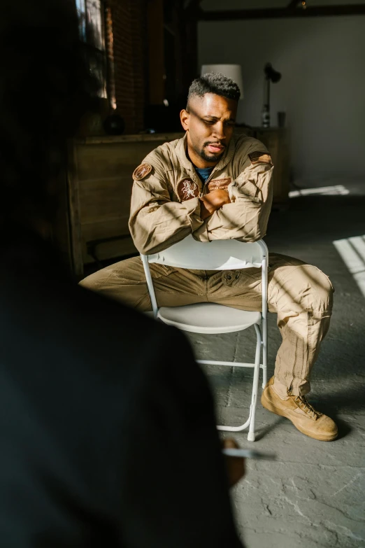 a man sitting on a chair in a room, air force jumpsuit, emotional conflict, tan suit, airforce gear