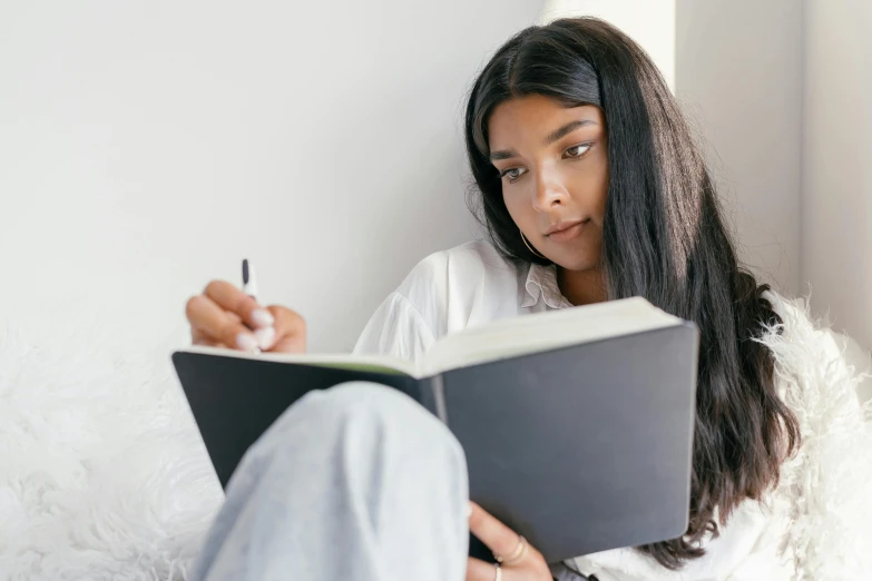 a woman sitting on a couch reading a book, a drawing, trending on pexels, female with long black hair, writing in journal, someone sits in bed, indian girl with brown skin