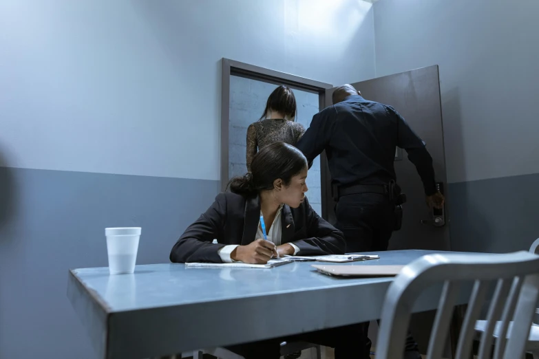 a woman sitting at a table in a room, police scene, profile image, prison scene, thumbnail