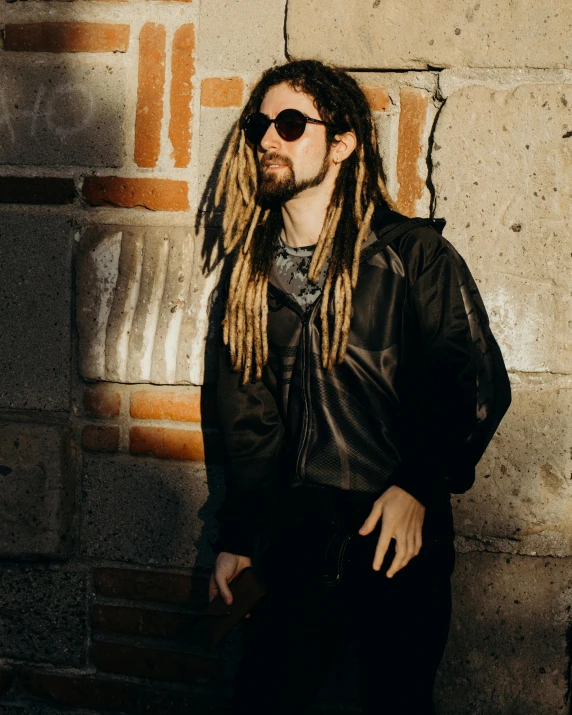 a man with long blonde hair and sunglasses standing next to a brick wall