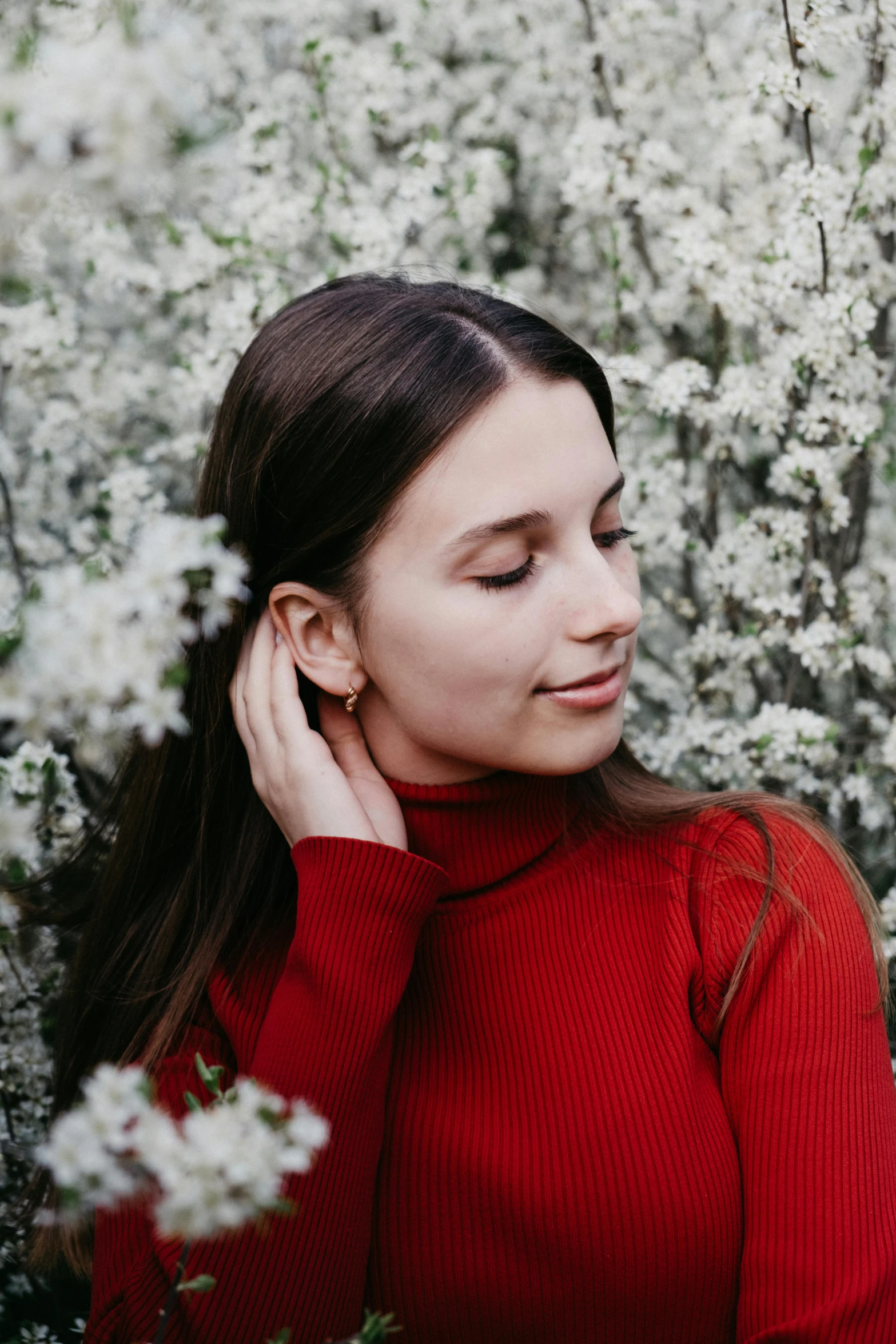 a woman standing in a field of white flowers, by Julia Pishtar, trending on pexels, wearing a red turtleneck sweater, earring, portrait sophie mudd, next to a tree