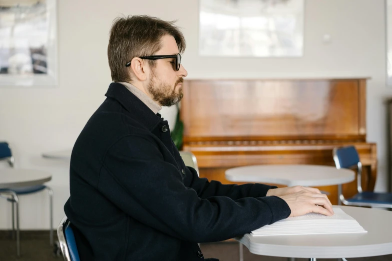 a man sitting at a table using a laptop computer, by Hallsteinn Sigurðsson, unsplash, private press, with glasses and goatee, bent - over posture, lachlan bailey, people sitting at tables