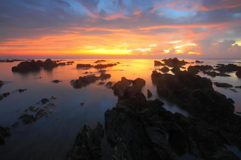 a sunset over a body of water with rocks in the foreground, new zeeland, image of the day, ultrawide shots, lava rock