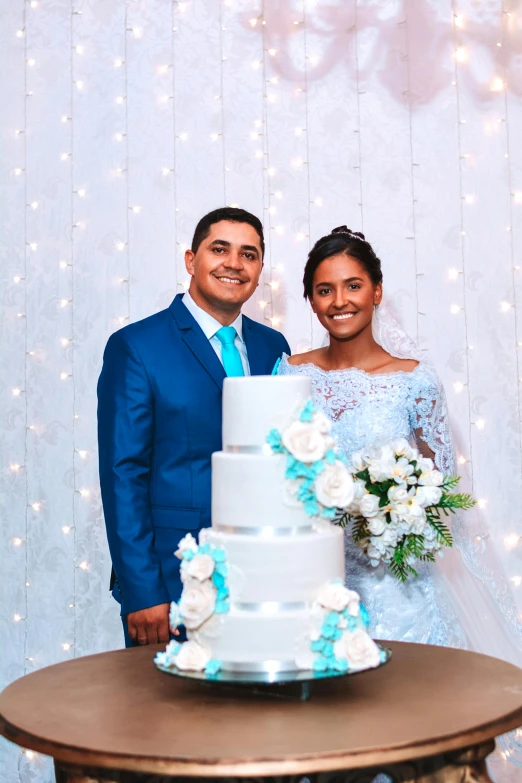 a bride and groom standing in front of a wedding cake, a colorized photo, by Robbie Trevino, pexels contest winner, wearing a light blue suit, alanis guillen, dazzling lights, in front of white back drop