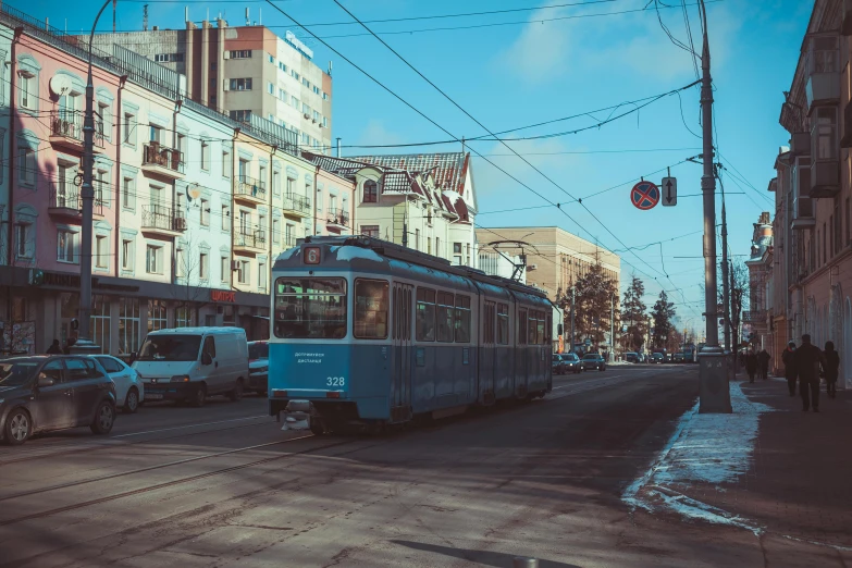 a blue train traveling down a street next to tall buildings, by Andrei Kolkoutine, pexels contest winner, dieselpunk volgograd, 🚿🗝📝, street tram, sunny winter day