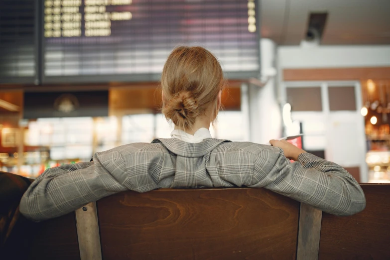 a woman sitting on a bench with her back to the camera, airport, sat at a desk, hair tied up in a bun, girl in a suit