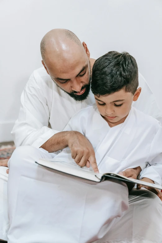 a man reading a book to a young boy, by Sheikh Hamdullah, pexels contest winner, hurufiyya, wearing white robe, gif, instruction, te pae