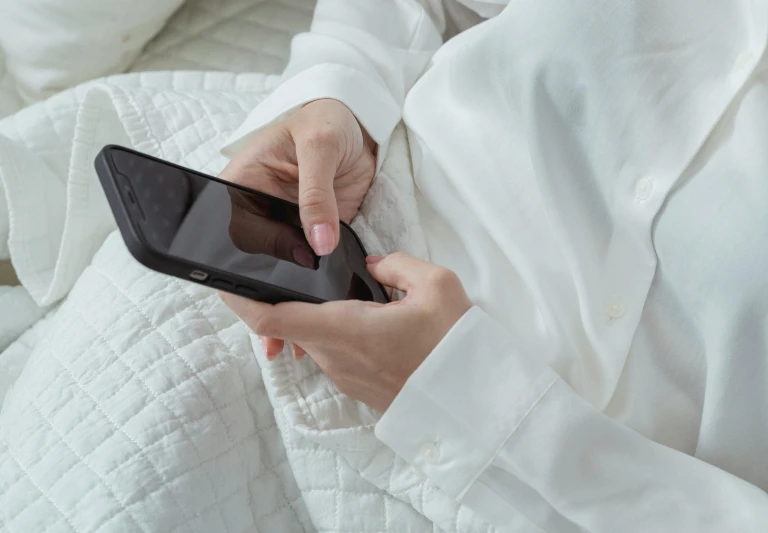 a close up of a person holding a cell phone, wearing white pajamas, sitting on the bed, white dress shirt, wearing a tracksuit