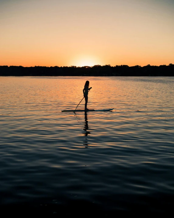 a person riding a paddle board on a body of water, in the evening, standing next to water