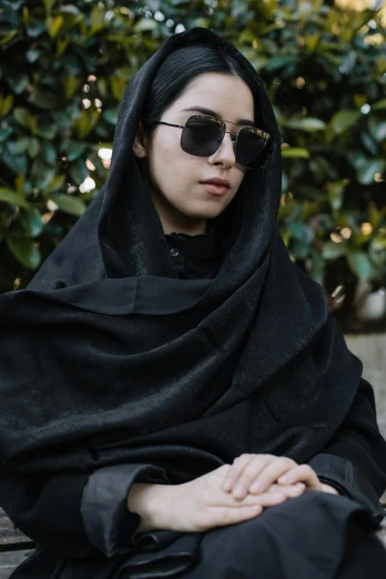 a woman sitting on a bench wearing a black shawl, an album cover, inspired by Maryam Hashemi, trending on unsplash, qajar art, wear ray - ban glass, black textured, androgynous person, vantablack gi