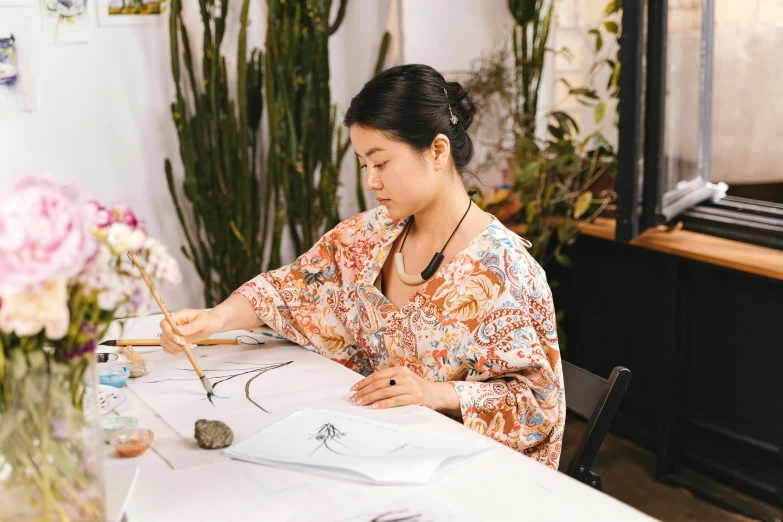 a woman sitting at a table writing on a piece of paper, inspired by Yun-Fei Ji, pexels contest winner, process art, ikebana, in a white boho style studio, wearing floral chiton, joy ang