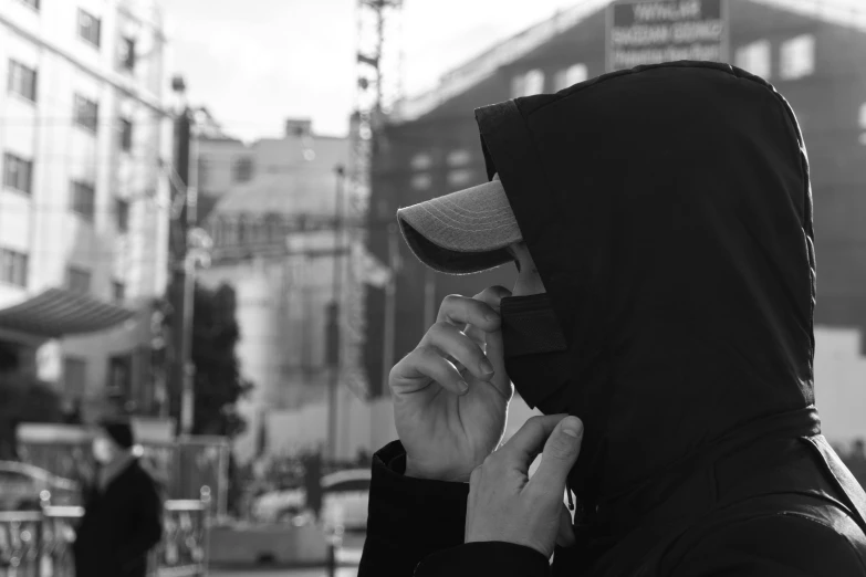 a man in a hood talking on a cell phone, a black and white photo, by Emma Andijewska, hand holding cap brim, black facemask, street pic, high quality upload