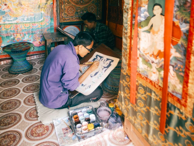 a man sitting on the floor in front of a painting, a detailed painting, pexels contest winner, cloisonnism, nepal, vibrant watercolor painting, a painting of two people, ornate designs on desk