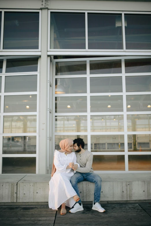 a man and woman sitting on a bench in front of a building, by Washington Allston, pexels contest winner, hurufiyya, white hijab, indoor picture, romantic lead, embracing