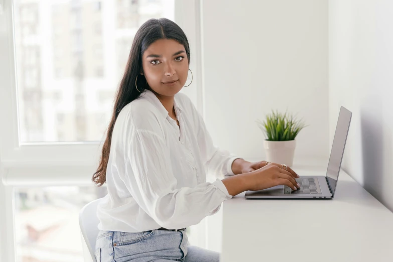 a woman sitting at a desk with a laptop, a character portrait, trending on pexels, indian girl with brown skin, dressed in white, in white room, young commoner