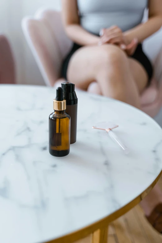 a woman sitting in a chair next to a table with a bottle of essential oil on it, by Will Ellis, trending on pexels, on a marble pedestal, brown resin, holding syringe, someone sits in bed