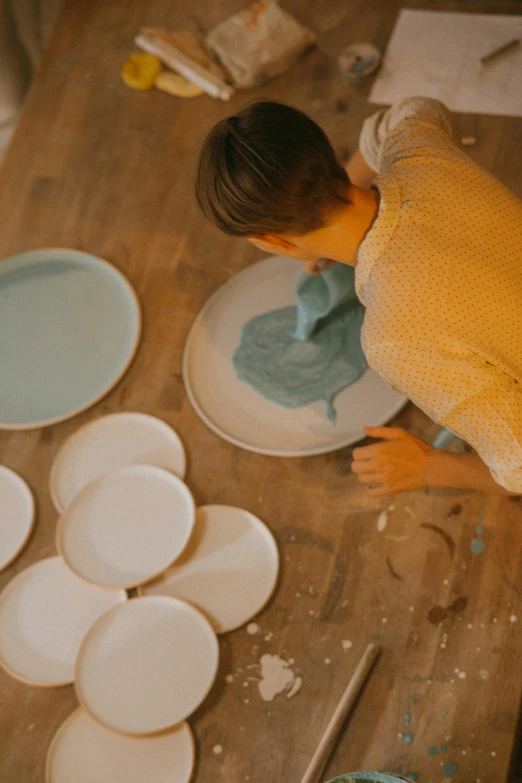 a woman is painting plates on a table, inspired by Constantin Hansen, trending on unsplash, with celadon glaze, big production, frank moth, facing away