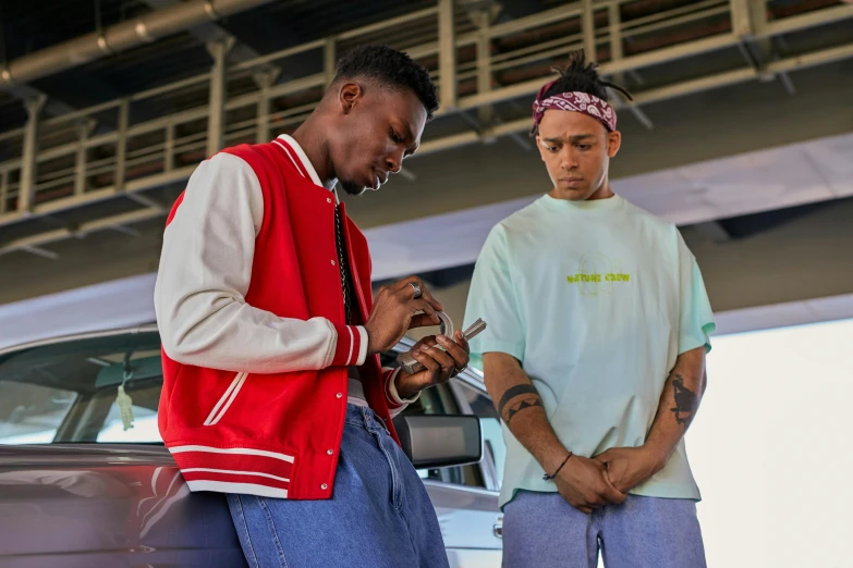 two men standing next to each other in front of a car, an album cover, trending on pexels, xxxtentacion, looking at his phone, wearing green clothing, wearing red and yellow clothes