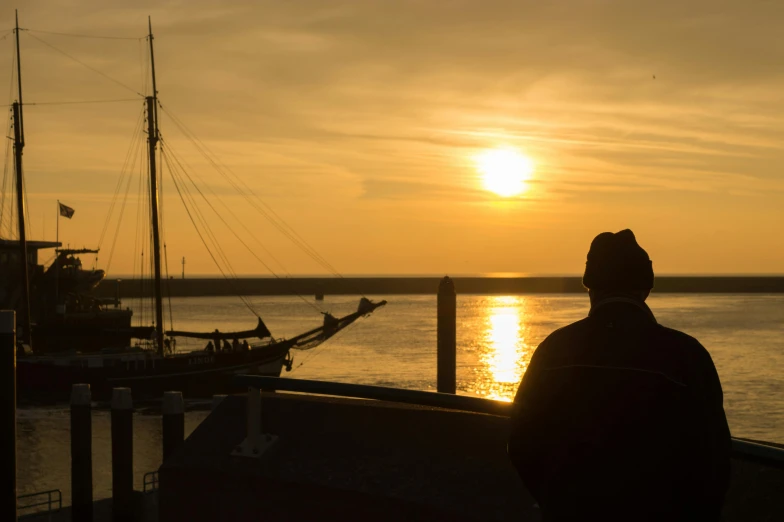 a person standing on a dock watching the sun set, by Jan Tengnagel, pexels contest winner, harbour, silhouette of a man, brown, maryport