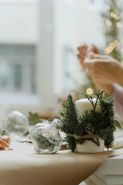 a woman sitting at a table in front of a laptop, organic ornaments, white candles, hands on counter, evergreen branches
