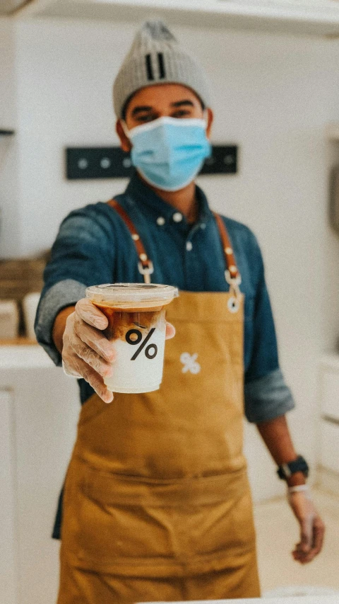 a man wearing a face mask holding a cup of coffee, trending on pexels, wearing an apron, promotional image, 64x64, exiting store