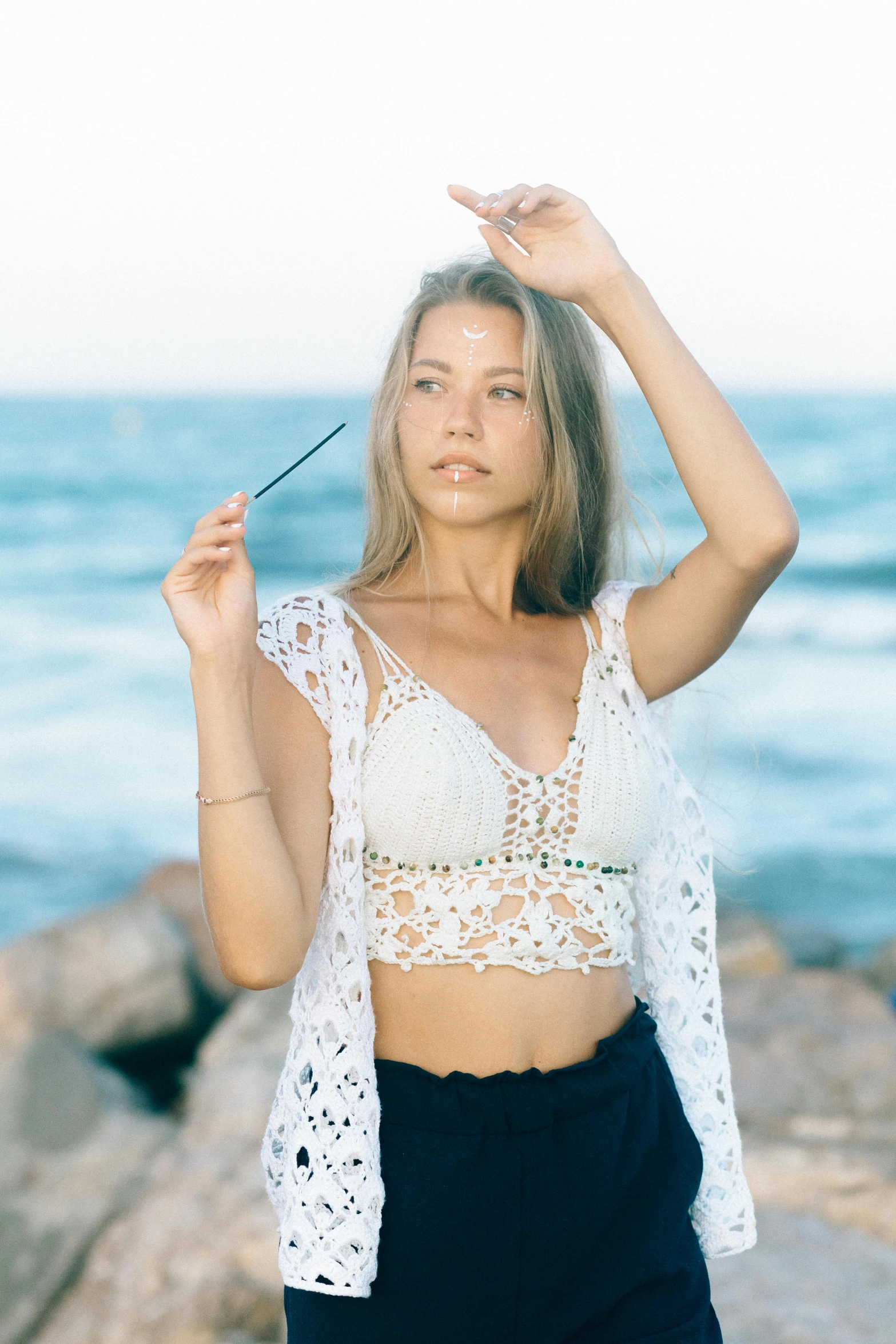 a beautiful young woman standing on top of a rocky beach, by Robbie Trevino, trending on pexels, renaissance, crochet, crop top, in a white boho style studio, model is wearing techtical vest