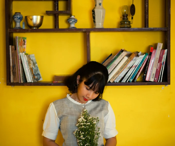 a woman holding a bouquet of flowers in front of a bookshelf, inspired by Ruth Jên, pexels contest winner, minimalism, yellow walls, vietnamese woman, avatar image, indoor shot