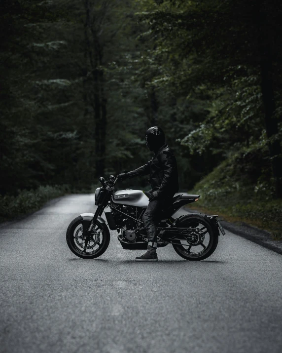 a man riding on the back of a motorcycle down a road, standing in a dark forest, wearing jeans and a black hoodie, man in silver space suit, top selection on unsplash