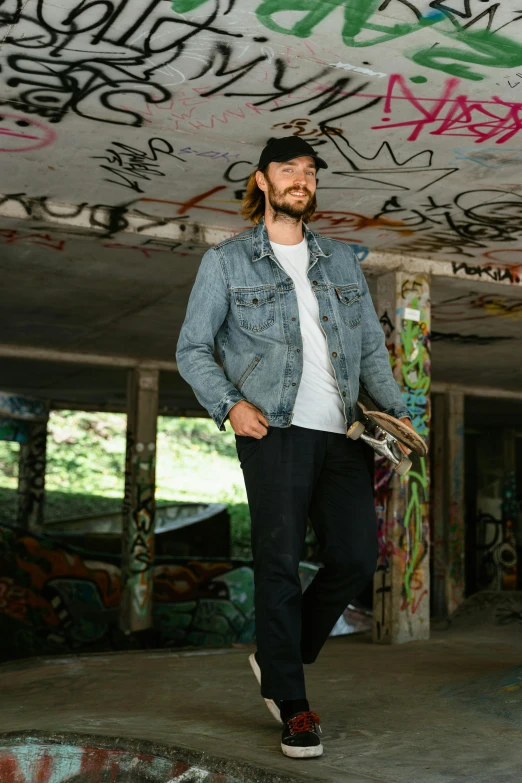 a man standing in a skate park holding a skateboard, a portrait, by Adam Dario Keel, wearing double denim, profile image, bearded, aura jared and wires