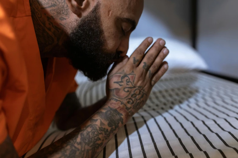 a man with tattoos is praying on a bed, trending on pexels, wearing orange prison jumpsuit, chris scalf, connection rituals, nipsey hussle